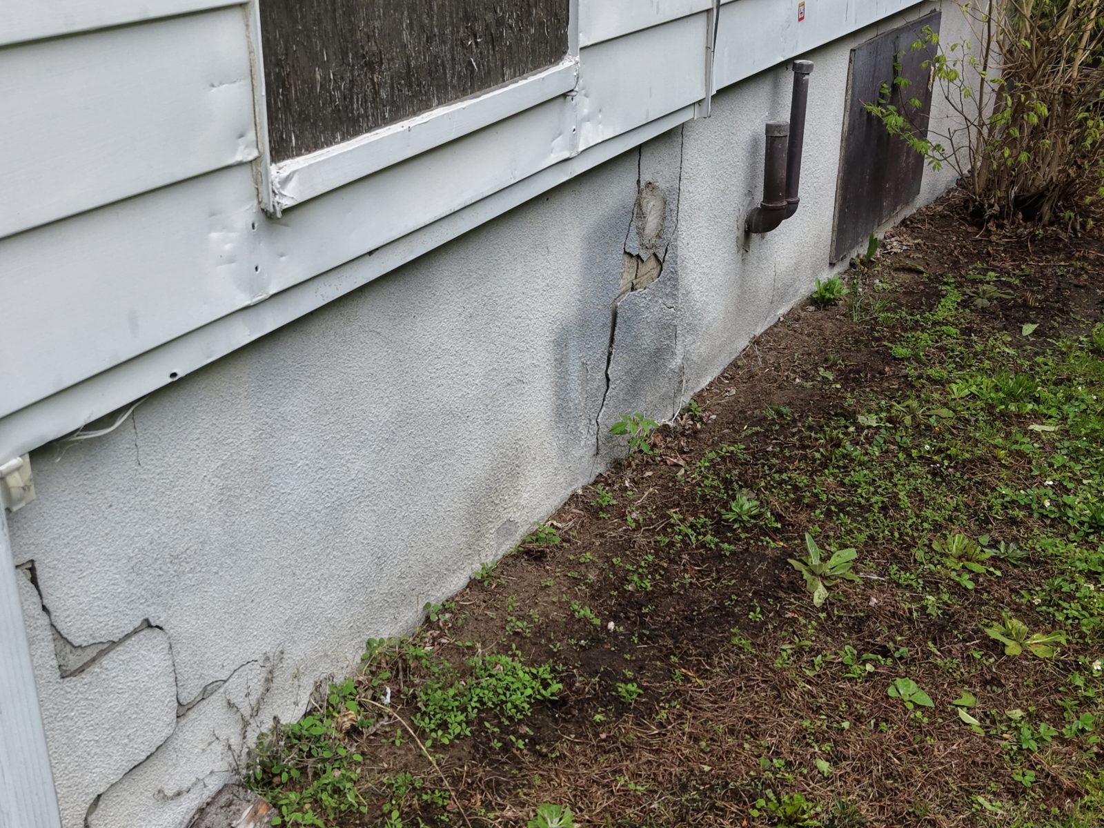 Crépi fissuré et manquant sur un mur de fondation en béton, avec une fissure verticale qui monte vers une fenêtre